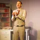 Mit eindringlichem Blick und gefalteten Händen spricht der Schauspieler direkt zum Publikum. Er steht vor einem hellen Hintergrund mit Bücherregal, warmes Licht fällt auf ihn – die Szene wirkt emotional und appellierend.