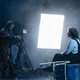 Eine Person wird im Scheinwerferlicht gefilmt, während eine andere die Kamera bedient. Die Szene ist von Dunkelheit, Nebel und Requisiten umgeben.