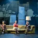Drei Personen sitzen auf einem Sofa auf der Bühne, mit Eis in der Hand und nostalgischer Kulisse im Hintergrund.