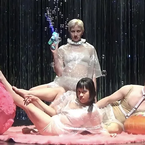 Drei Personen in transparenter Kleidung posieren auf einer Bühne mit Glitzer-Vorhang. Sie sitzen zwischen Plüschball, Donut-Schwimmring und Seifenblasen – ein surrealer, performativer Szenenaufbau voller Ironie.