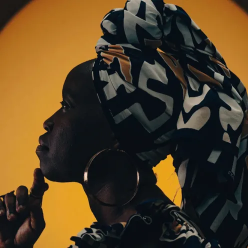 Frau im Profil vor gelbem Hintergrund, mit farbig gemustertem Kopftuch und großen Creolen. Sie wirkt nachdenklich, ihr Finger ruht am Kinn. Das Licht betont Silhouette und Ausdruck auf eindrucksvolle Weise.