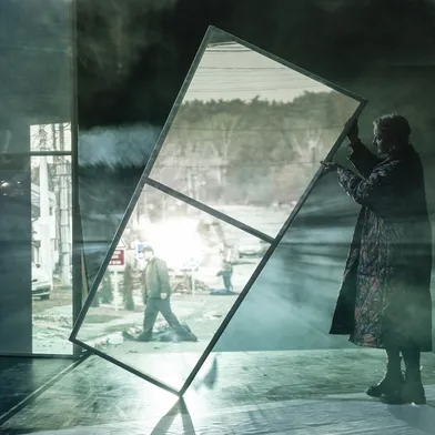 Eine Frau hält einen schrägen Rahmen auf der Bühne, während im Hintergrund eine Kriegslandschaft projiziert wird.