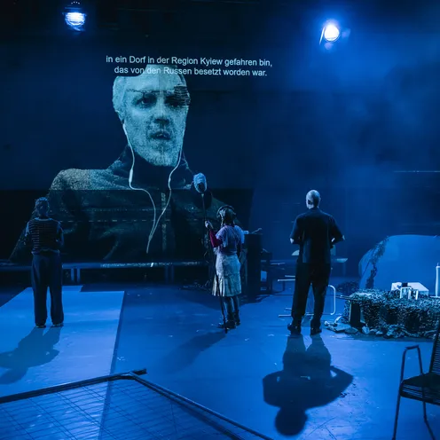 Drei Personen stehen vor einer großen Video-Projektion eines Mannes mit Untertiteln über den Krieg in der Ukraine. Die Szene ist blau beleuchtet.