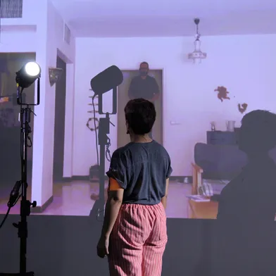 Eine Frau steht vor einer Projektion, in der ein Mann im Türrahmen eines Wohnzimmers zu sehen ist. Zwei Studiolichter werfen Schatten auf die Leinwand. Die Szene wirkt wie eine künstlerische Performance oder Theaterinszenierung.