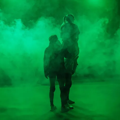 Zwei Frauen mit heller Haut stehen Rücken an Rücken in dichtem, grünem Nebel. Ihre Silhouetten sind sichtbar, beide tragen sportliche Kleidung und Turnschuhe. Das Licht ist grün und schafft eine geheimnisvolle, dramatische Atmosphäre.