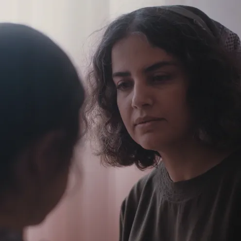 Eine Frau mit nachdenklichem Blick steht einem Kind gegenüber, das nur von hinten zu sehen ist. Die Szene wirkt ruhig und emotional, mit weichem Licht und zarten Farben im Hintergrund.