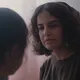 Eine Frau mit nachdenklichem Blick steht einem Kind gegenüber, das nur von hinten zu sehen ist. Die Szene wirkt ruhig und emotional, mit weichem Licht und zarten Farben im Hintergrund.