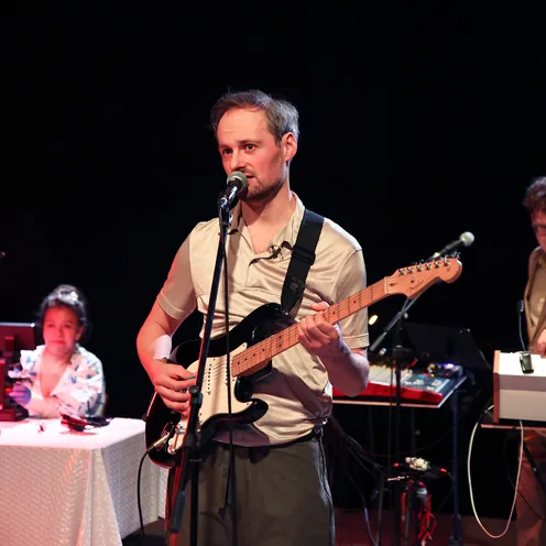 Drei Personen stehen auf einer Bühne: Ein Mann mit heller Haut singt und spielt E-Gitarre. Im Hintergrund bedient ein weiterer Mann mit heller Haut ein Keyboard. Links sitzt eine Frau mit mittlerem Hautton an einem Tisch mit Technik und blickt zur Bühne.