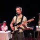 Drei Personen stehen auf einer Bühne: Ein Mann mit heller Haut singt und spielt E-Gitarre. Im Hintergrund bedient ein weiterer Mann mit heller Haut ein Keyboard. Links sitzt eine Frau mit mittlerem Hautton an einem Tisch mit Technik und blickt zur Bühne.