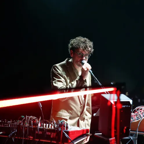 Ein Mann mit heller Haut spricht oder singt an einem Mikrofon neben elektronischen Musikinstrumenten und Kabeln. Er trägt ein helles Sakko und eine Brille. Eine rote Lichtleiste verläuft quer durch das Bild und verstärkt die Bühnenatmosphäre.