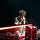 Ein Mann mit heller Haut spricht oder singt an einem Mikrofon neben elektronischen Musikinstrumenten und Kabeln. Er trägt ein helles Sakko und eine Brille. Eine rote Lichtleiste verläuft quer durch das Bild und verstärkt die Bühnenatmosphäre.