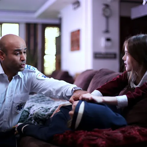 Ein Sanitäter betreut ein krankes Kind auf einem Sofa, während ein Mädchen besorgt dessen Hand hält. Die Szene spielt in einem Wohnzimmer mit warmem Licht und wirkt fürsorglich und ruhig.