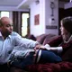 Ein Sanitäter betreut ein krankes Kind auf einem Sofa, während ein Mädchen besorgt dessen Hand hält. Die Szene spielt in einem Wohnzimmer mit warmem Licht und wirkt fürsorglich und ruhig.