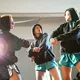 Drei Personen in schwarzen Hoodies und kurzen Sporthosen tanzen synchron in einem Raum. Ein grelles Licht im Hintergrund erzeugt eine dramatische Atmosphäre, während alle konzentriert in eine Richtung blicken.