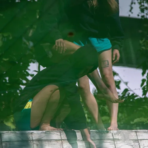 Drei Personen in Hoodies stehen barfuß an einem leeren, mit Fliesen ausgekleideten Pool. Ihre Spiegelung im Wasser und das grüne Licht schaffen eine surreale, fast träumerische Szene. Eine Hand ragt aus dem Wasser.