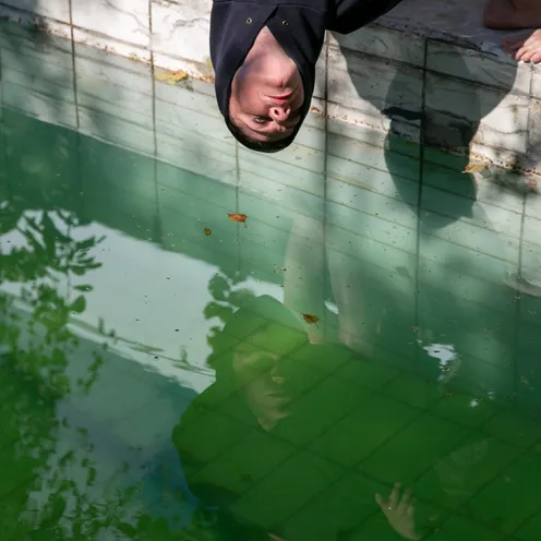Eine Person lehnt kopfüber über den Beckenrand eines grünen Pools, das Gesicht spiegelt sich im Wasser. Die Szene wirkt ruhig und introspektiv, mit weichem Licht und herbstlichen Blättern auf dem Boden.