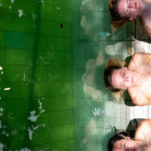 Drei Personen liegen mit dem Kopf am Beckenrand eines grünen Pools. Ihre Körper spiegeln sich im Wasser. Der Ausdruck reicht von entspannt bis wachsam, während Licht und Wasser Reflexionen erzeugen.
