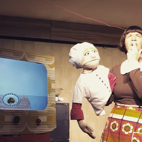 Eine Frau mit brauner Perücke und bunter Schürze steht neben einer Handpuppe in Kochkleidung. Die Szene wirkt wie aus einer Retro-Kochshow mit Fernsehapparat und Humor.