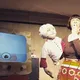 Eine Frau mit brauner Perücke und bunter Schürze steht neben einer Handpuppe in Kochkleidung. Die Szene wirkt wie aus einer Retro-Kochshow mit Fernsehapparat und Humor.