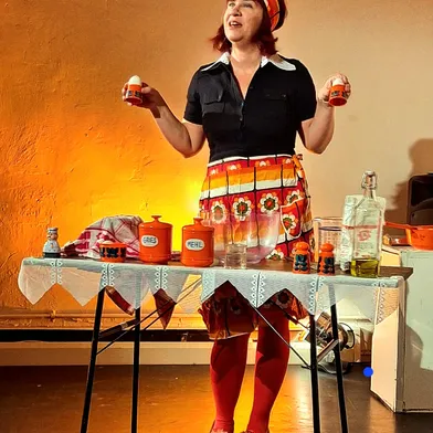 Eine Frau in bunter Retro-Kleidung steht an einem gedeckten Tisch mit Gewürzen und Zutaten. Sie lächelt und hält zwei Dosen in den Händen, als würde sie ein nostalgisches Kochprogramm präsentieren.