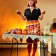 Eine Frau in bunter Retro-Kleidung steht an einem gedeckten Tisch mit Gewürzen und Zutaten. Sie lächelt und hält zwei Dosen in den Händen, als würde sie ein nostalgisches Kochprogramm präsentieren.