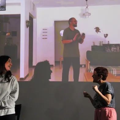 Zwei Frauen stehen lächelnd vor einer Projektion, die einen Mann in einem Wohnzimmer zeigt. Beide tragen Mikrofone, was auf eine Bühnenperformance oder interaktive Präsentation hindeutet. Die Stimmung wirkt lebendig und gespannt.