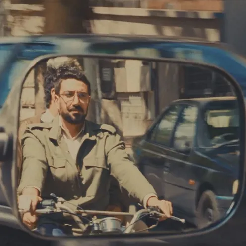 Ein Mann mit Brille und Bart fährt Motorrad, sein Spiegelbild ist in einem Rückspiegel eines Autos zu sehen. Die Szene spielt in einer belebten Stadtstraße bei Tageslicht und wirkt filmisch inszeniert.