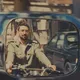 Ein Mann mit Brille und Bart fährt Motorrad, sein Spiegelbild ist in einem Rückspiegel eines Autos zu sehen. Die Szene spielt in einer belebten Stadtstraße bei Tageslicht und wirkt filmisch inszeniert.