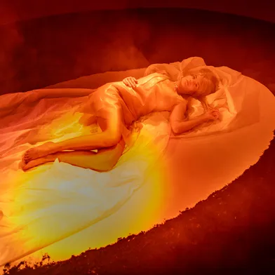Eine Frau liegt in einem orange beleuchteten Kreis auf weißen Tüchern. Ihr Körper ist in ein seidenartiges Kleid gehüllt, der Raum wirkt neblig und geheimnisvoll. Die Lichtstimmung erinnert an Feuer oder Sonnenaufgang.