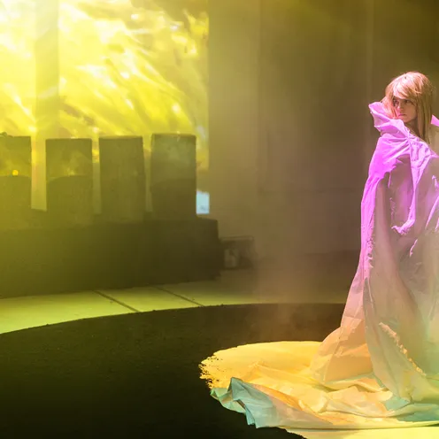 Die Frau steht nun im gleichen Kreis, in ein weißes Tuch gehüllt. Gelbes Licht fällt auf sie, im Hintergrund sind Projektionen und Baumstämme zu sehen. Die Szene wirkt traumartig und entrückt.