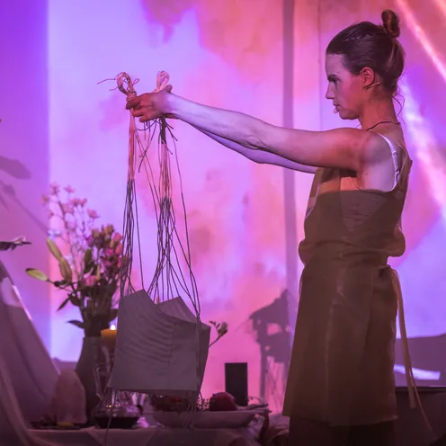 Eine Frau hält entschlossen ein Objekt mit langen Fäden vor sich. Sie steht vor einem Altar mit Blumen, Kerzen und Früchten, in magischem, rosa-violettem Licht. Eine rituelle Handlung scheint im Gange zu sein.