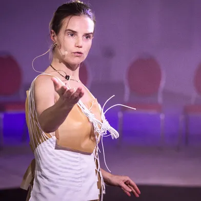 Die Frau spricht mit ausdrucksstarker Geste, trägt ein kunstvolles Kleid und ein Mikrofon. Im Hintergrund stehen leere rote Stühle, die Szenerie erinnert an ein performatives Theaterstück oder eine Performance.