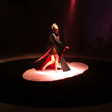 Die Frau tanzt in einem schwarzen Umhang mit rotem Innenfutter, barfuß auf einem hell beleuchteten Kreis in einem dunklen Raum. Ihre Bewegung ist dynamisch und kraftvoll – eine tänzerische Ausdrucksform.