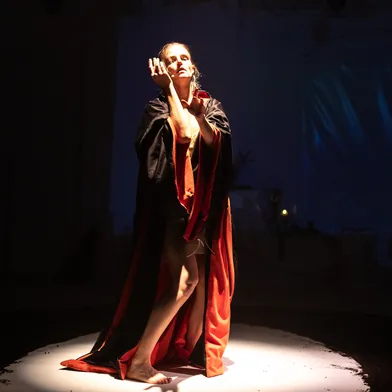 Im Schein eines Spotlights posiert die Frau in einem dramatischen Moment, den Blick erhoben, die Arme anmutig geführt. Der Umhang öffnet sich, Licht und Schatten erzeugen eine sakrale, theatralische Stimmung.