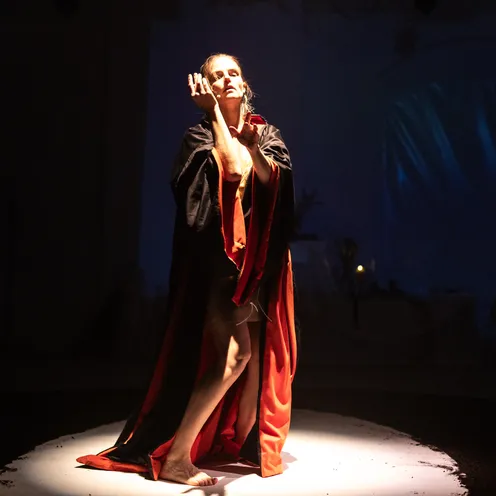 Im Schein eines Spotlights posiert die Frau in einem dramatischen Moment, den Blick erhoben, die Arme anmutig geführt. Der Umhang öffnet sich, Licht und Schatten erzeugen eine sakrale, theatralische Stimmung.
