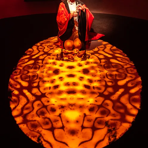 Eine Frau liegt in einem orange beleuchteten Kreis auf weißen Tüchern. Ihr Körper ist in ein seidenartiges Kleid gehüllt, der Raum wirkt neblig und geheimnisvoll. Die Lichtstimmung erinnert an Feuer oder Sonnenaufgang.