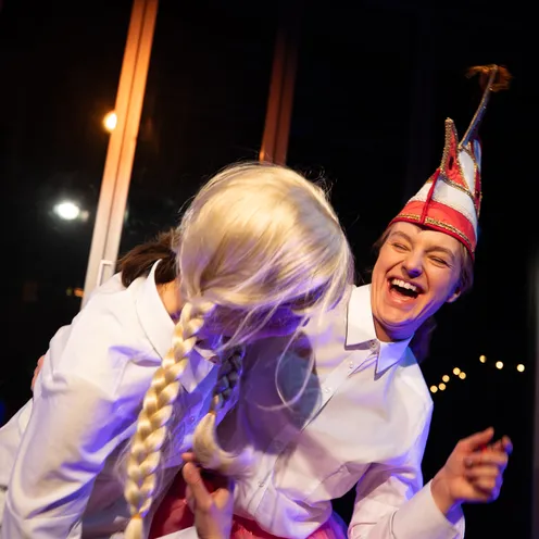 Zwei Frauen in weißem Hemd und dunkler Hose machen einen synchronen Tanzschritt auf einer Bühne. Sie lachen und tragen gestreifte Socken – die Atmosphäre ist ausgelassen und kabarettistisch.