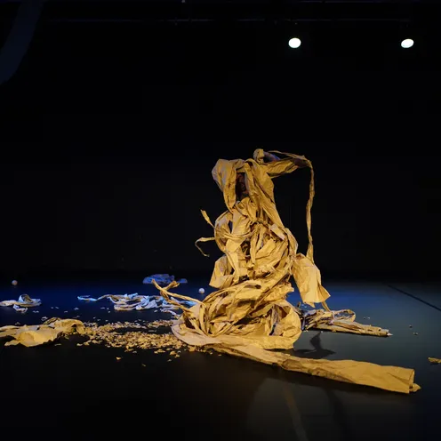 Auf dunkler Bühne liegt ein chaotischer Haufen aus zerknülltem, braunem Papier, teilweise aufgerichtet wie eine Skulptur. Einzelne Papierstreifen und Holzkugeln sind verstreut, Spotlight setzt die Szene dramatisch in Szene.