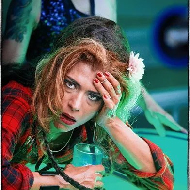 Eine Frau mit zerzausten Haaren, rotem Karohemd und Blume im Haar stützt sich erschöpft mit einem Drink auf einen Tisch. Ihr Blick ist intensiv, fast verzweifelt – ein Moment zwischen Rausch, Müdigkeit und Emotion.