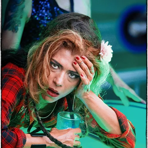 Eine Frau mit zerzausten Haaren, rotem Karohemd und Blume im Haar stützt sich erschöpft mit einem Drink auf einen Tisch. Ihr Blick ist intensiv, fast verzweifelt – ein Moment zwischen Rausch, Müdigkeit und Emotion.