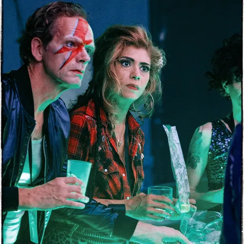 Eine Frau im Karohemd und ein Mann mit David-Bowie-Blitz im Gesicht schauen fassungslos auf etwas vor ihnen. Sie halten Getränke und Eisbeutel in den Händen, die Szene wirkt angespannt und surreal – ein stiller Höhepunkt der Geschichte.