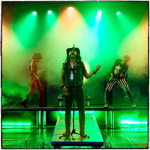 Drei Musiker stehen auf einer in grünes Licht getauchten Bühne, umgeben von Nebel. Der Mann in der Mitte singt leidenschaftlich ins Mikro, während die anderen in rockiger Pose mitspielen – ein kraftvoller Moment im Konzert.