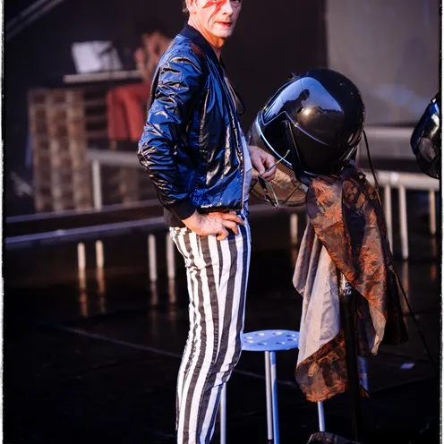 Ein Mann mit Bowie-Gesichtsbemalung steht selbstbewusst auf einer Bühne, gestreifte Hose und silberne Schuhe unterstreichen seinen auffälligen Look. Im Hintergrund eine Umkleideszene – der Moment wirkt wie vor oder nach einem Auftritt.