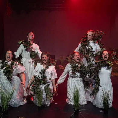 Sechs Personen auf einer Bühne, überwiegend Frauen und ein Mann mit heller Haut, tragen weiße Gewänder und sind mit grünen Pflanzen geschmückt. Sie knien und schreien dramatisch, rot beleuchtet, mit intensiven Gesichtsausdrücken in einer theatralischen Szene.