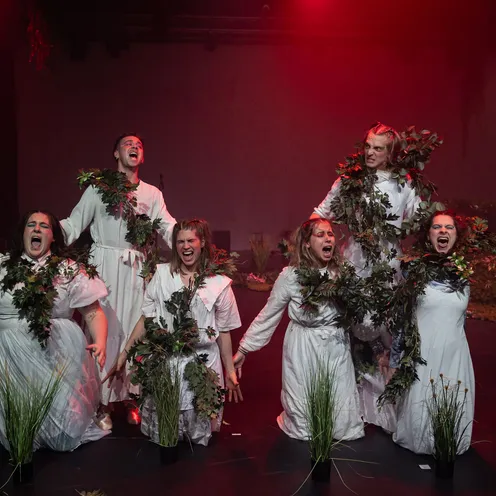Sechs Personen auf einer Bühne, überwiegend Frauen und ein Mann mit heller Haut, tragen weiße Gewänder und sind mit grünen Pflanzen geschmückt. Sie knien und schreien dramatisch, rot beleuchtet, mit intensiven Gesichtsausdrücken in einer theatralischen Szene.