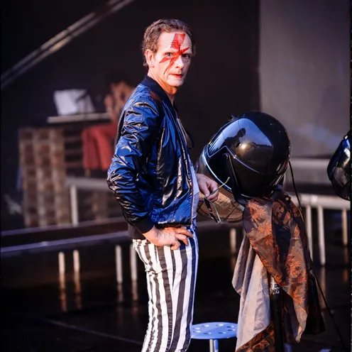 Ein Mann mit Bowie-Gesichtsbemalung steht selbstbewusst auf einer Bühne, gestreifte Hose und silberne Schuhe unterstreichen seinen auffälligen Look. Im Hintergrund eine Umkleideszene – der Moment wirkt wie vor oder nach einem Auftritt.