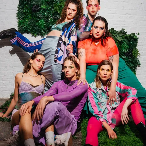 Sechs Personen posieren expressiv vor einer Wand mit grüner Pflanzen-Deko. Bunte, auffällige Kleidung, Make-up und intensive Blicke betonen Individualität und künstlerischen Ausdruck. Die Szene wirkt modisch, mutig und kreativ.