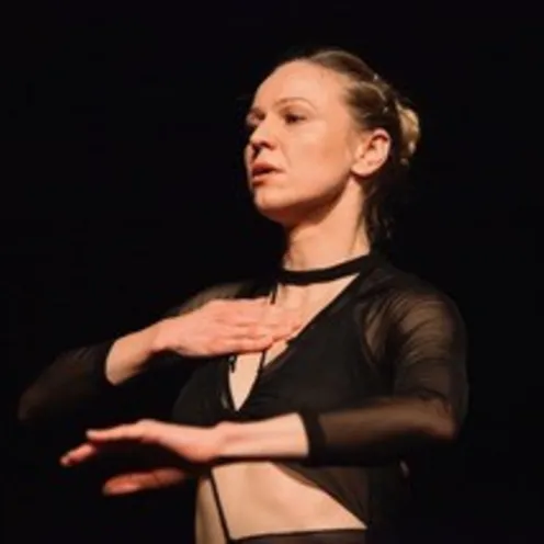 Eine Tänzerin in schwarzem Outfit steht vor dunklem Hintergrund. Mit geschlossenen Augen und ausdrucksstarker Geste legt sie eine Hand auf die Brust, die andere ist erhoben – ein Moment voller Konzentration und Emotion.