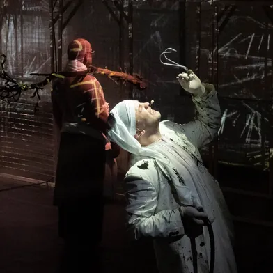 Ein weißhäutiger Mann in weißem Kostüm hebt einen metallenen Haken, sein Gesicht dramatisch beleuchtet. Im Hintergrund steht eine zweite Person, vermutlich eine Frau mit dunkler Haut, in einem rot-gemusterten Kostüm.