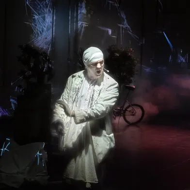 Ein weißhäutiger Mann in weißem Kostüm und Haube steht auf einer düsteren Bühne, gestikuliert dramatisch und trägt ein Headset. Hinter ihm sind Nebel, Pflanzen und ein Fahrrad zu sehen, die Szene wirkt theatralisch und surreal.